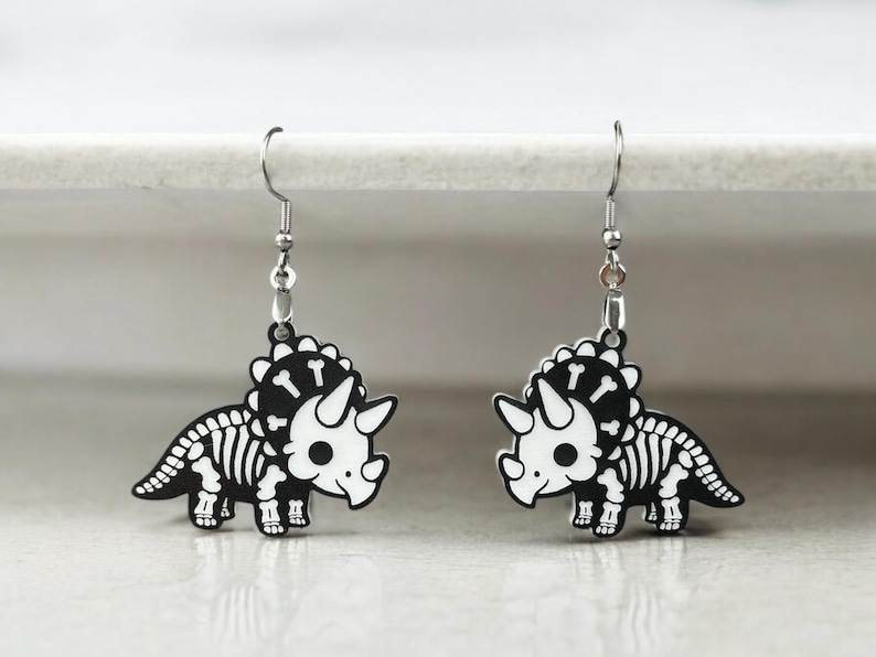 Puede incluir: Un par de pendientes de plata con un dise&ntilde;o de triceratops esqueleto en blanco y negro. Los pendientes est&aacute;n hechos de metal y tienen un dise&ntilde;o colgante.