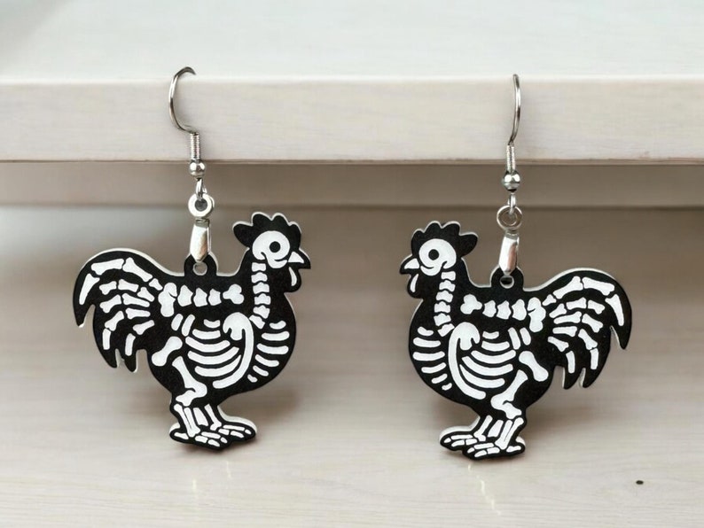 Puede incluir: Un par de pendientes de pollo esqueleto de plata. Los esqueletos de pollo son negros y blancos y tienen un dise&ntilde;o detallado.