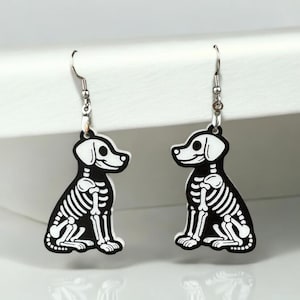 Puede incluir: Un par de pendientes de plata con colgantes de perro esqueleto en blanco y negro. Los perros est&aacute;n sentados con la cola enroscada.