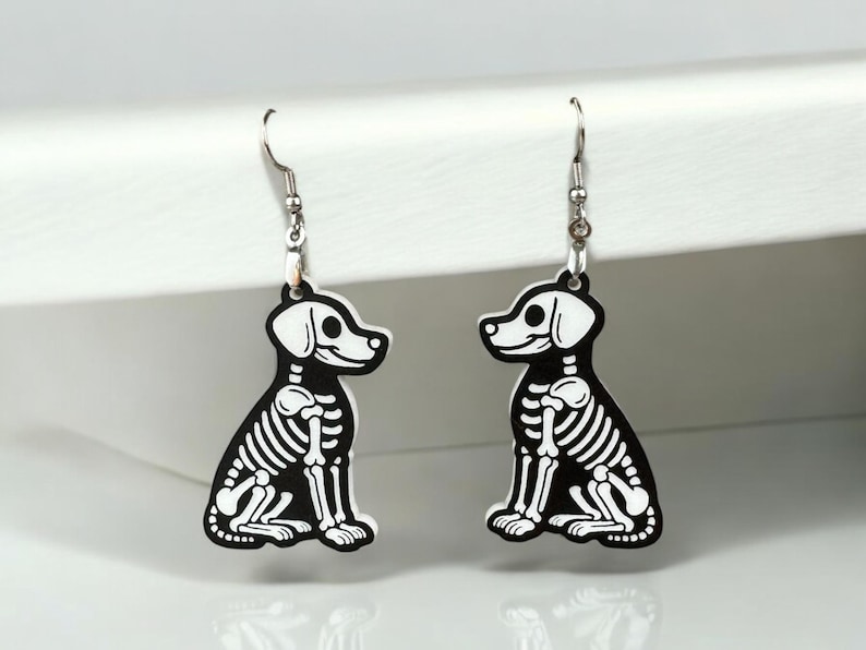 Puede incluir: Un par de pendientes de plata con colgantes de perro esqueleto en blanco y negro. Los perros est&aacute;n sentados con la cola enroscada.