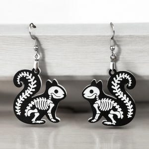 Puede incluir: Un par de pendientes de ardilla esqueleto en blanco y negro. Los pendientes est&aacute;n hechos de metal y tienen un dise&ntilde;o colgante.