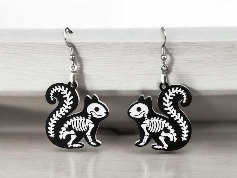 Puede incluir: Un par de pendientes de ardilla esqueleto en blanco y negro. Los pendientes est&aacute;n hechos de metal y tienen un dise&ntilde;o colgante.