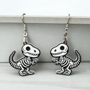 Puede incluir: Un par de pendientes de plata con forma de esqueleto de dinosaurio. Los dinosaurios son blancos con contornos negros y est&aacute;n en posici&oacute;n de caminar.