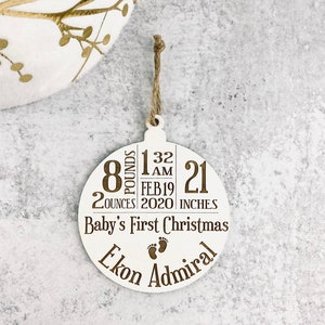 K&ouml;nnte beinhalten: Wei&szlig;er Weihnachtsbaumschmuck aus Holz mit einer Juteschnur. Der Schmuck hat den eingravierten Text "Baby's First Christmas" und enth&auml;lt die Geburtsdaten des Babys: 3,6 kg, 53 cm, und den Namen "Ekon Admiral".