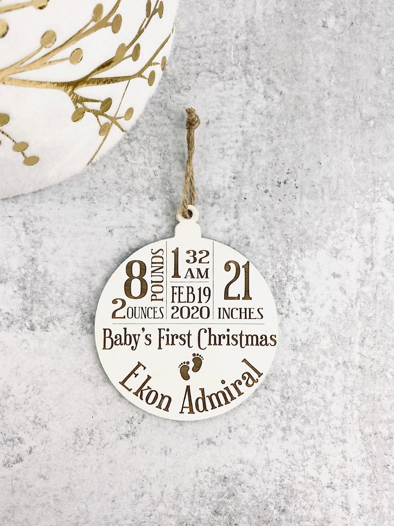 K&ouml;nnte beinhalten: Wei&szlig;er Weihnachtsbaumschmuck aus Holz mit einer Juteschnur. Der Schmuck hat den eingravierten Text "Baby's First Christmas" und enth&auml;lt die Geburtsdaten des Babys: 3,6 kg, 53 cm, und den Namen "Ekon Admiral".