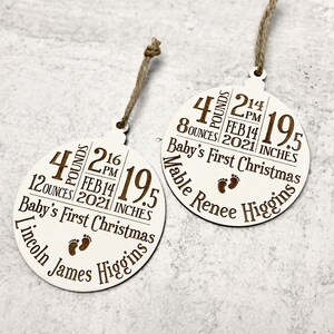 K&ouml;nnte beinhalten: Zwei wei&szlig;e runde Holz-Weihnachtsornamente mit braunem Text und Kordelaufh&auml;ngern. Jedes Ornament zeigt die Geburtsdaten des Babys: Gewicht in Pfund und Unzen, Uhrzeit, Datum, L&auml;nge in Zoll und den Namen des Babys. Der Text "Baby's First Christmas" ist ebenfalls enthalten.