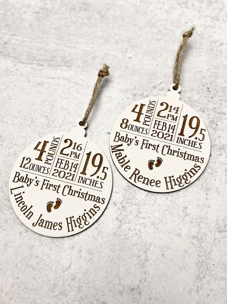 K&ouml;nnte beinhalten: Zwei wei&szlig;e runde Holz-Weihnachtsornamente mit braunem Text und Kordelaufh&auml;ngern. Jedes Ornament zeigt die Geburtsdaten des Babys: Gewicht in Pfund und Unzen, Uhrzeit, Datum, L&auml;nge in Zoll und den Namen des Babys. Der Text "Baby's First Christmas" ist ebenfalls enthalten.
