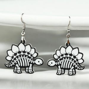 Puede incluir: Un par de pendientes de plata con un dise&ntilde;o de esqueleto de dinosaurio blanco. El dinosaurio tiene una espalda espinosa y una cara linda.
