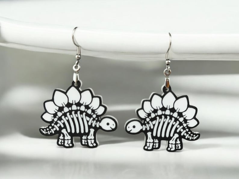 Puede incluir: Un par de pendientes de plata con un dise&ntilde;o de esqueleto de dinosaurio blanco. El dinosaurio tiene una espalda espinosa y una cara linda.