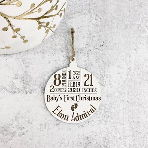 K&ouml;nnte beinhalten: Ein wei&szlig;er, kreisf&ouml;rmiger Weihnachtsschmuck mit braunem Text und einer Schnurschlaufe. Der Schmuck zeigt die Geburtsdaten des Babys: 3,6 kg, 56,6 g, 53,34 cm, geboren am 19. Februar 2020, um 1 Uhr morgens. Er tr&auml;gt die Aufschrift "Baby's First Christmas" und den Namen "Ekon Admiral".