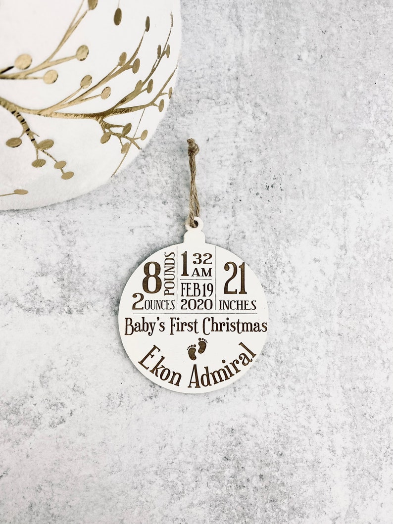 K&ouml;nnte beinhalten: Ein wei&szlig;er, kreisf&ouml;rmiger Weihnachtsschmuck mit braunem Text und einer Schnurschlaufe. Der Schmuck zeigt die Geburtsdaten des Babys: 3,6 kg, 56,6 g, 53,34 cm, geboren am 19. Februar 2020, um 1 Uhr morgens. Er tr&auml;gt die Aufschrift "Baby's First Christmas" und den Namen "Ekon Admiral".