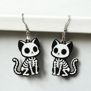 Puede incluir: Un par de pendientes de aro de plata con dijes de gato esqueleto en blanco y negro. Los gatos est&aacute;n sentados con sus colas enroscadas alrededor de sus patas.