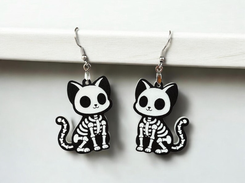 Puede incluir: Un par de pendientes de aro de plata con dijes de gato esqueleto en blanco y negro. Los gatos est&aacute;n sentados con sus colas enroscadas alrededor de sus patas.