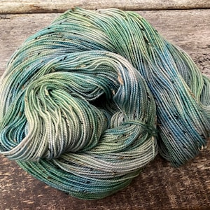 May include: A skein of hand-dyed yarn in shades of teal, green, and blue with speckled accents. The yarn is tightly wound, showcasing its texture and color variations. Ideal for knitting or crocheting projects.