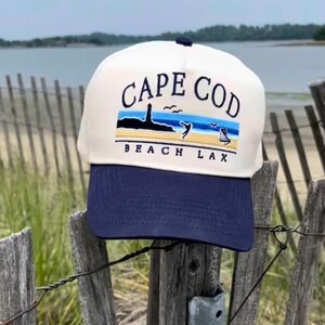 Puede incluir: Gorra de béisbol blanca y azul marino con el texto "CAPE COD BEACH LAX" bordado en la parte delantera. La gorra presenta una escena de playa con un faro, olas y un jugador de lacrosse. La gorra está sobre una valla de madera.