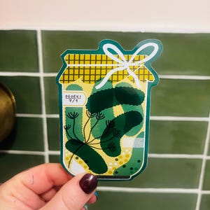 May include: A sticker depicting a jar of pickles, labelled "OGORKI 7/9". The jar has a yellow, checkered lid and a white bow. Inside are green pickles and dill. The sticker has a green border.