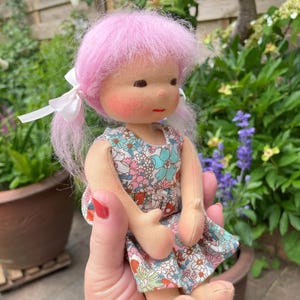 May include: A handmade doll with pink yarn hair styled in pigtails with white bows. The doll has rosy cheeks, a small red mouth, and is wearing a floral print dress. The doll is made of fabric and has a whimsical, handcrafted appearance.