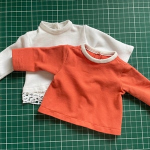 Puede incluir: Dos camisas de manga larga, una blanca con un borde de encaje y una naranja.