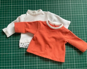 Puede incluir: Dos camisas de manga larga, una blanca con un borde de encaje y una naranja.
