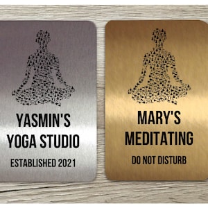 Può includere: Due cartelli in metallo argento e oro con una posa di yoga fatta di pose di yoga più piccole. Il cartello argentato dice "Yasmin's Yoga Studio Established 2021". Il cartello dorato dice "Mary's Meditating Do Not Disturb".