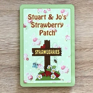 Puede incluir: Un cartel verde con borde blanco y un patrón floral. El cartel dice "Stuart & Jo's Strawberry Patch" y tiene una cruz de madera con un cartel que dice "Strawberries".