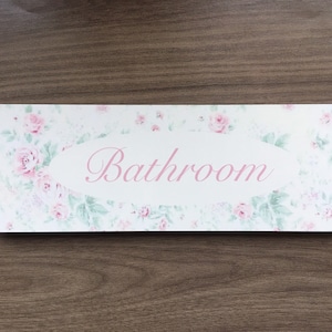 Puede incluir: Un letrero blanco con un patrón floral rosa y la palabra "Baño" en letras rosas.