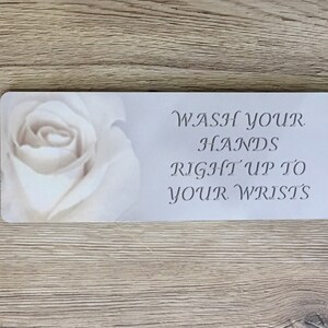 May include: White sign with a white rose image and the text "WASH YOUR HANDS RIGHT UP TO YOUR WRISTS".