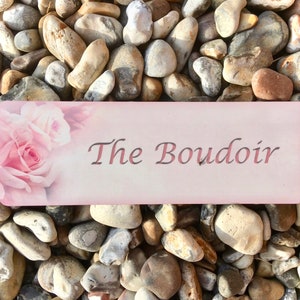 May include: A pink and white sign with the text "The Boudoir" in a cursive font. The sign is decorated with a pink rose and has a light pink background.