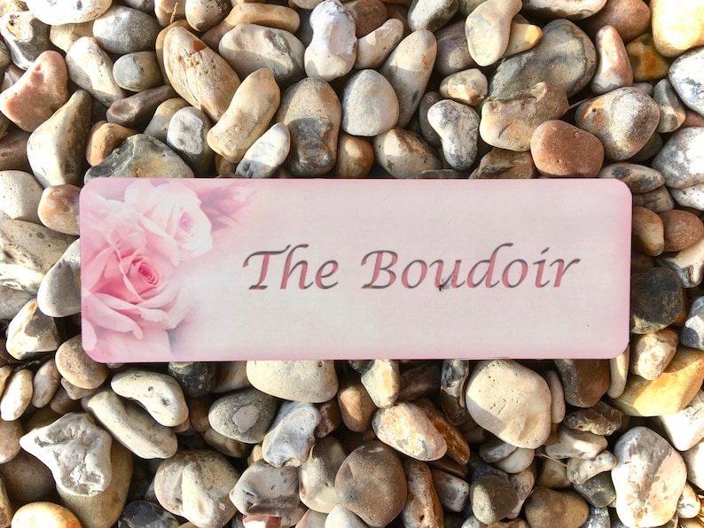 May include: A pink and white sign with the text "The Boudoir" in a cursive font. The sign is decorated with a pink rose and has a light pink background.