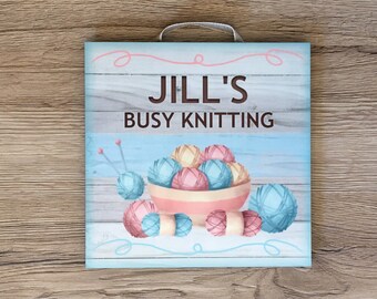 May include: A blue and white wooden sign with the text "Jill's Busy Knitting" and a cartoon image of a bowl of yarn balls.