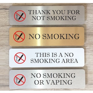 May include: Four silver and gold metal signs with a no smoking symbol and text. The signs read "Thank you for not smoking", "No smoking", "This is a no smoking area", and "No smoking or vaping".