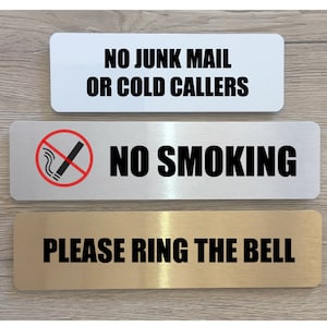 May include: Three silver and gold signs with black text. The top sign says "No Junk Mail or Cold Callers". The middle sign says "No Smoking" with a red circle and a diagonal line through a cigarette. The bottom sign says "Please Ring The Bell".