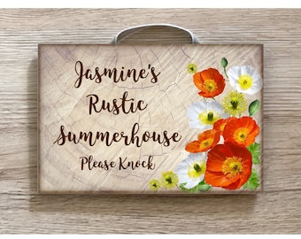 Könnte beinhalten: Ein rustikales Holzschild mit dem Text "Jasmine's Rustic Summerhouse Please Knock" in braunen Buchstaben. Das Schild ist mit einem floralen Design verziert, das orange und weiße Mohnblumen zeigt.