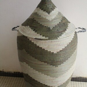 May include: A tall, woven basket with a conical lid. The basket features a repeating pattern of gray, white, and black horizontal stripes. It has two small handles on the sides and a knob on the lid. The basket is made of natural fibers.