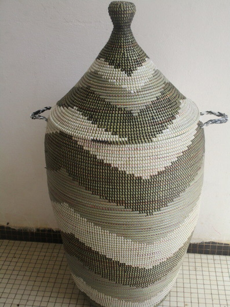 May include: A tall, woven basket with a conical lid. The basket features a repeating pattern of gray, white, and black horizontal stripes. It has two small handles on the sides and a knob on the lid. The basket is made of natural fibers.