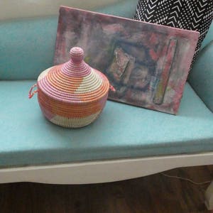 May include: A pink, orange and white woven basket with a lid sits on a light blue sofa. The basket is made from natural materials and has a unique, handcrafted look.