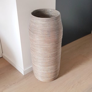 Op de afbeelding: Een hoge, cilindrische geweven mand in neutrale tinten. De mand is gemaakt van natuurlijke vezels, met horizontale banden van lichte en donkere kleuren. Hij staat op een lichte houten vloer, vlakbij een witte muur en een donkergrijze muur.