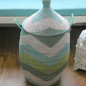 May include: A large, woven storage basket with a lid. The basket is made of natural materials and has a light blue, green, and white pattern. The basket is sitting on a wooden floor.