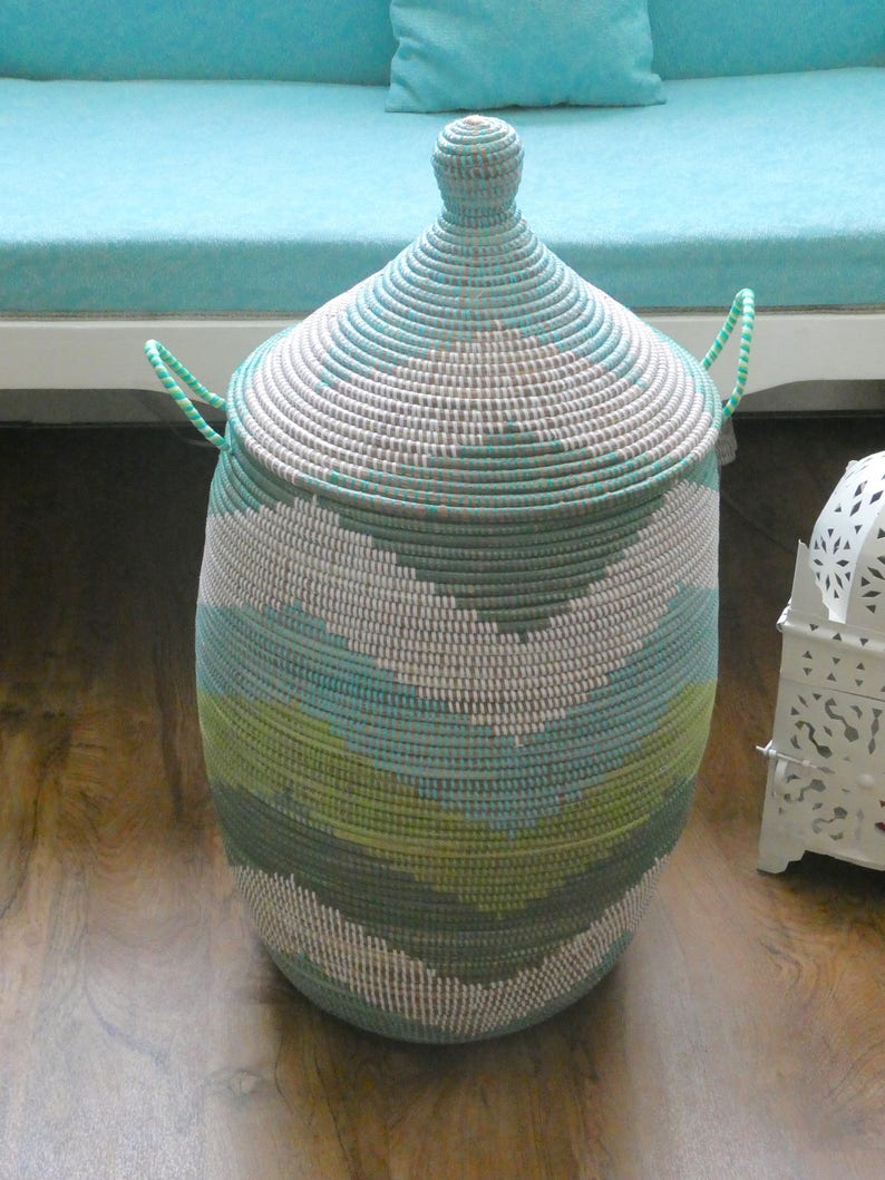 May include: A large, woven storage basket with a lid. The basket is made of natural materials and has a light blue, green, and white pattern. The basket is sitting on a wooden floor.