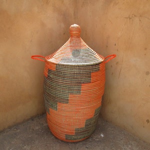 May include: A large, woven storage basket with a lid. The basket is made of natural materials and has a geometric pattern in shades of orange and green.