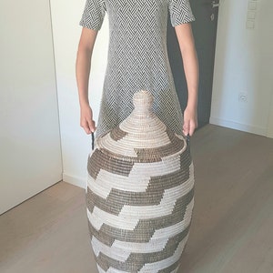 May include: A large, woven storage basket with a lid. The basket features a geometric pattern in shades of brown and white. The basket is held by a person wearing a black and white patterned dress.