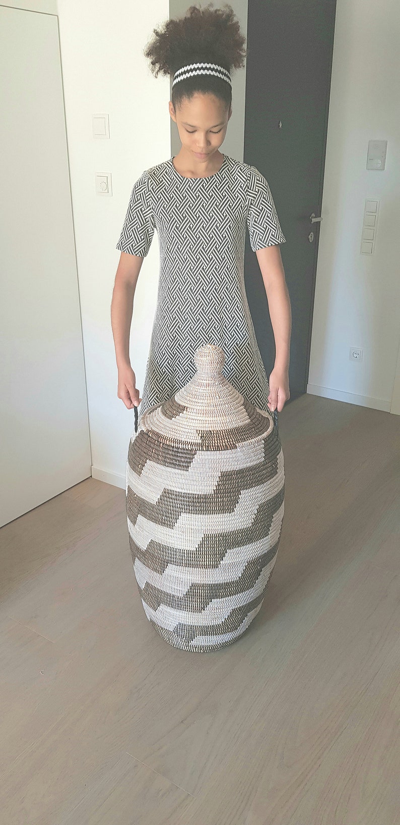 May include: A large, woven storage basket with a lid. The basket features a geometric pattern in shades of brown and white. The basket is held by a person wearing a black and white patterned dress.