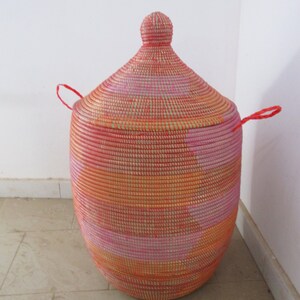 May include: A large, round, woven storage basket with a lid. The basket is made of natural fibres and has a pink, orange, and white colour scheme. The lid has a small knob on top and two red handles.