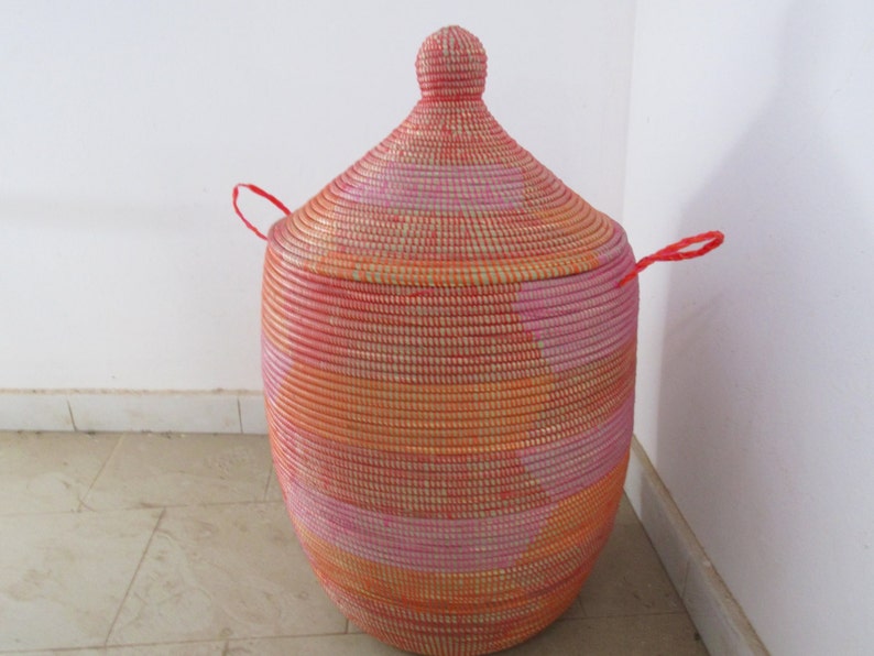 May include: A large, round, woven storage basket with a lid. The basket is made of natural fibres and has a pink, orange, and white colour scheme. The lid has a small knob on top and two red handles.