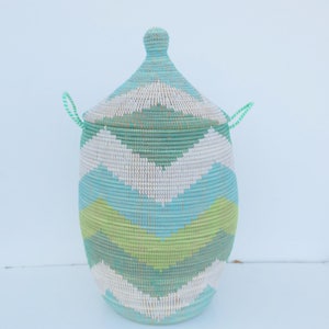 May include: A woven storage basket with a lid, featuring a geometric pattern in shades of green, blue, and white. The basket has two handles on the lid.