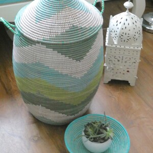 May include: A woven storage basket with a light blue, green, and white pattern. The basket has a lid and two handles. A small white planter with green succulents sits on a woven tray with a turquoise blue color.