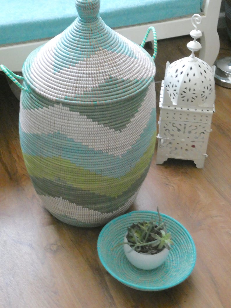 May include: A woven storage basket with a light blue, green, and white pattern. The basket has a lid and two handles. A small white planter with green succulents sits on a woven tray with a turquoise blue color.