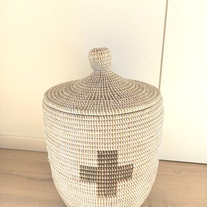 May include: A white and brown woven storage basket with a lid. The basket has a large, brown plus sign design on the front.