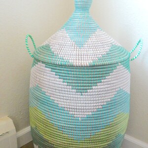 May include: A large, woven storage basket with a lid. The basket is made of natural materials and is painted in a geometric pattern of white, green, and blue.
