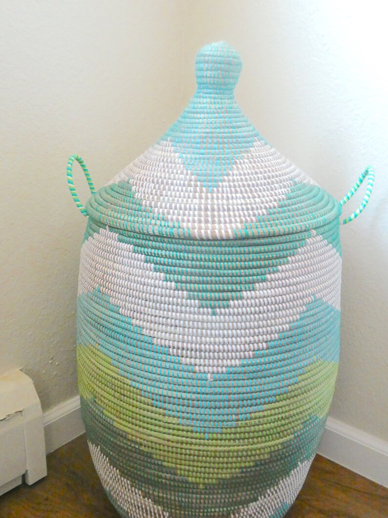 May include: A large, woven storage basket with a lid. The basket is made of natural materials and is painted in a geometric pattern of white, green, and blue.